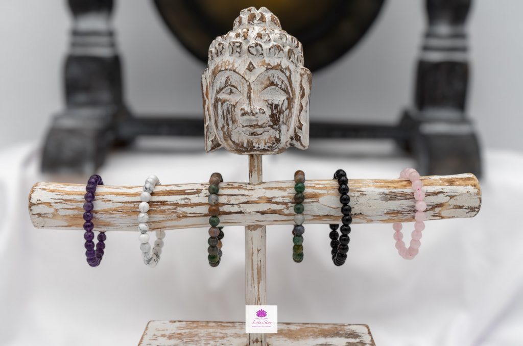 Wooden Buddha head sculpture with five beaded bracelets in purple, white, green, black, and pink, displayed on a rustic wooden stand.